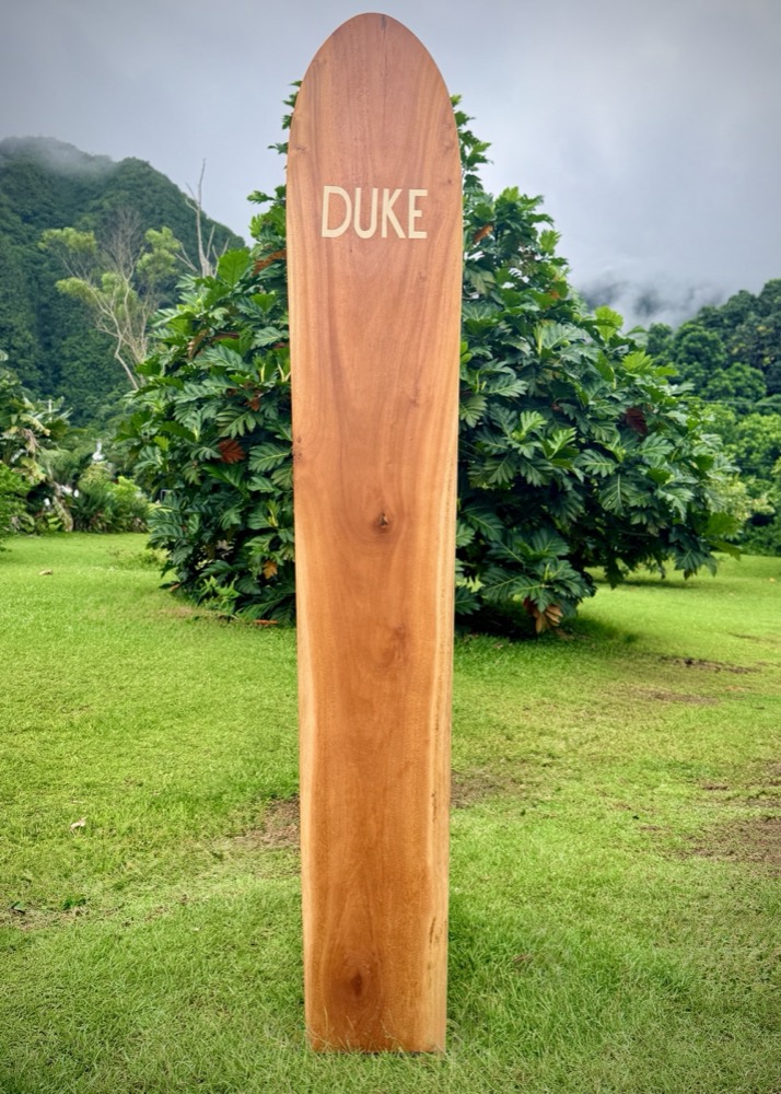 A wooden surfboard standing upright on its tail in mown grass, honey brown with a narrow oval nose and a few dark knots. The name DUKE is inlaid near the nose in a pale wood. Breadfruit trees behind it.